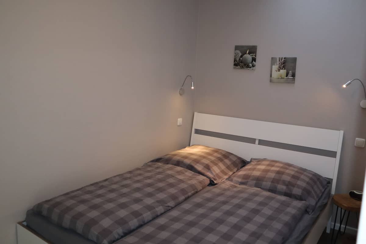 A single bed with a checkered bedspread and two pillows, a bedside table with a lamp, and two wall-mounted pictures.