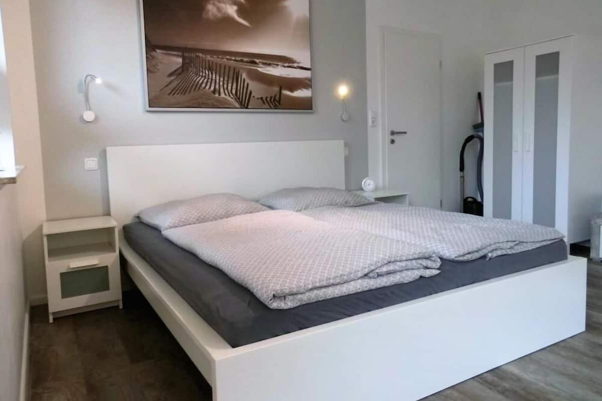 A modern bedroom with a large bed, a nightstand, a wall-mounted lamp, and a door with a vacuum cleaner.