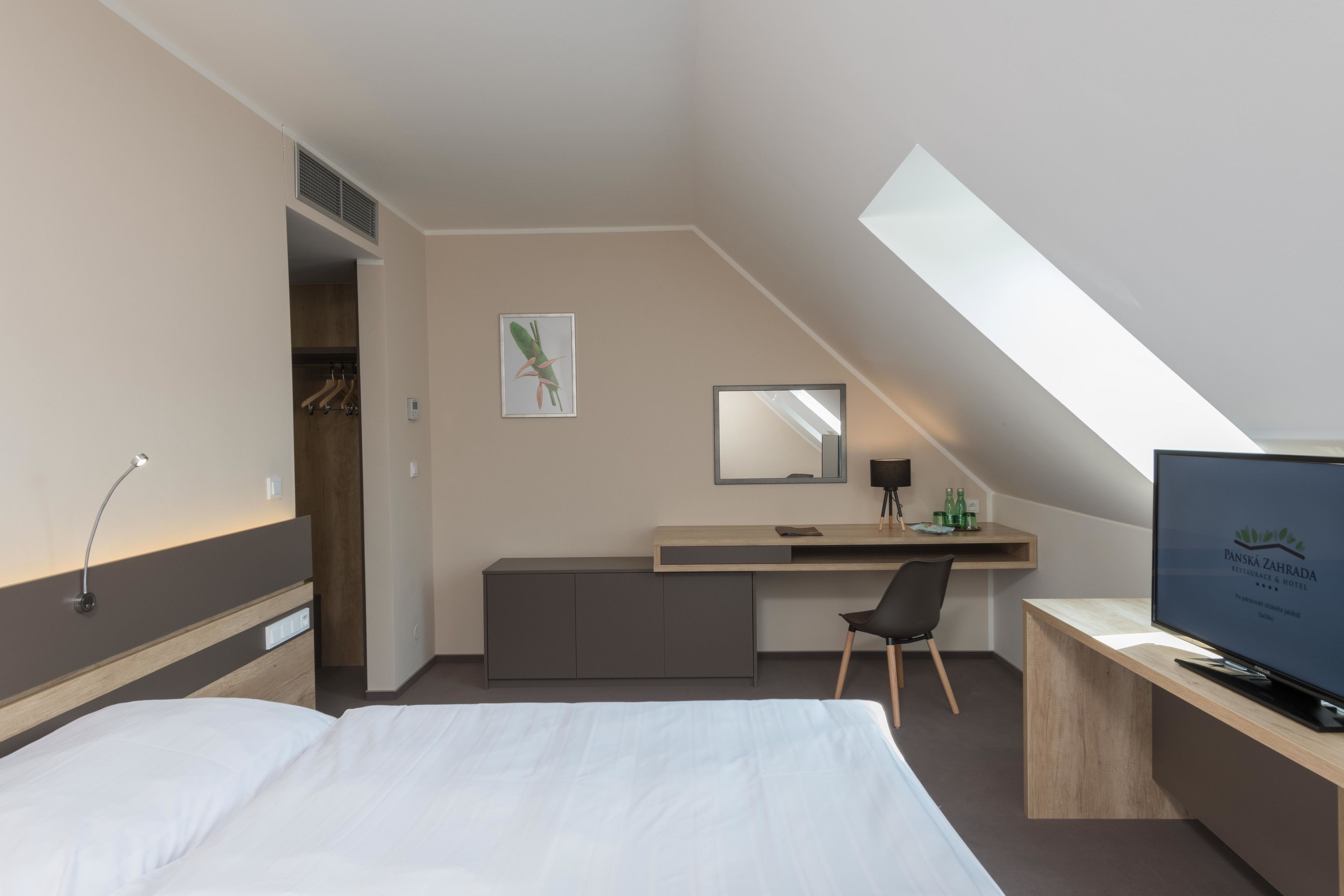 A modern hotel room with a bed, a desk, a chair, a television, and a skylight.