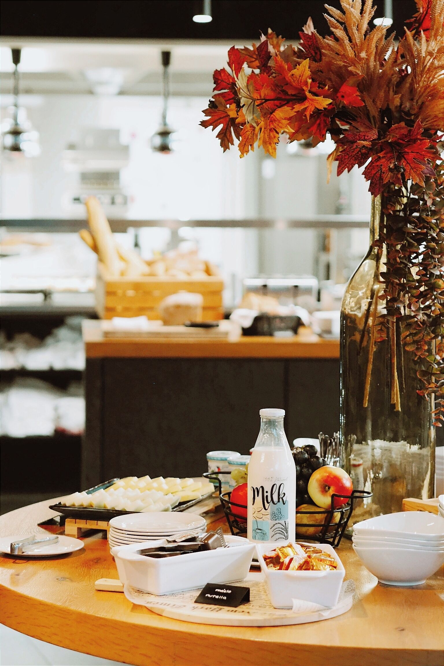 Une table de petit-déjeuner avec une bouteille de lait, des assiettes, des bols et un vase rempli de feuilles d’automne.