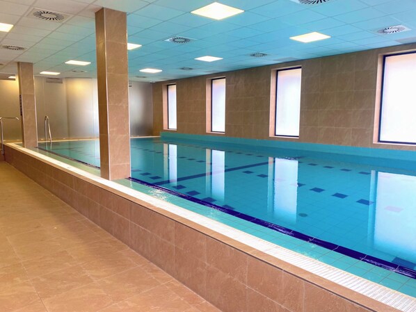 A long indoor swimming pool with clear blue water, surrounded by walls with windows and a tiled floor. - Hotel Panská Zahrada (Dobrichovice)