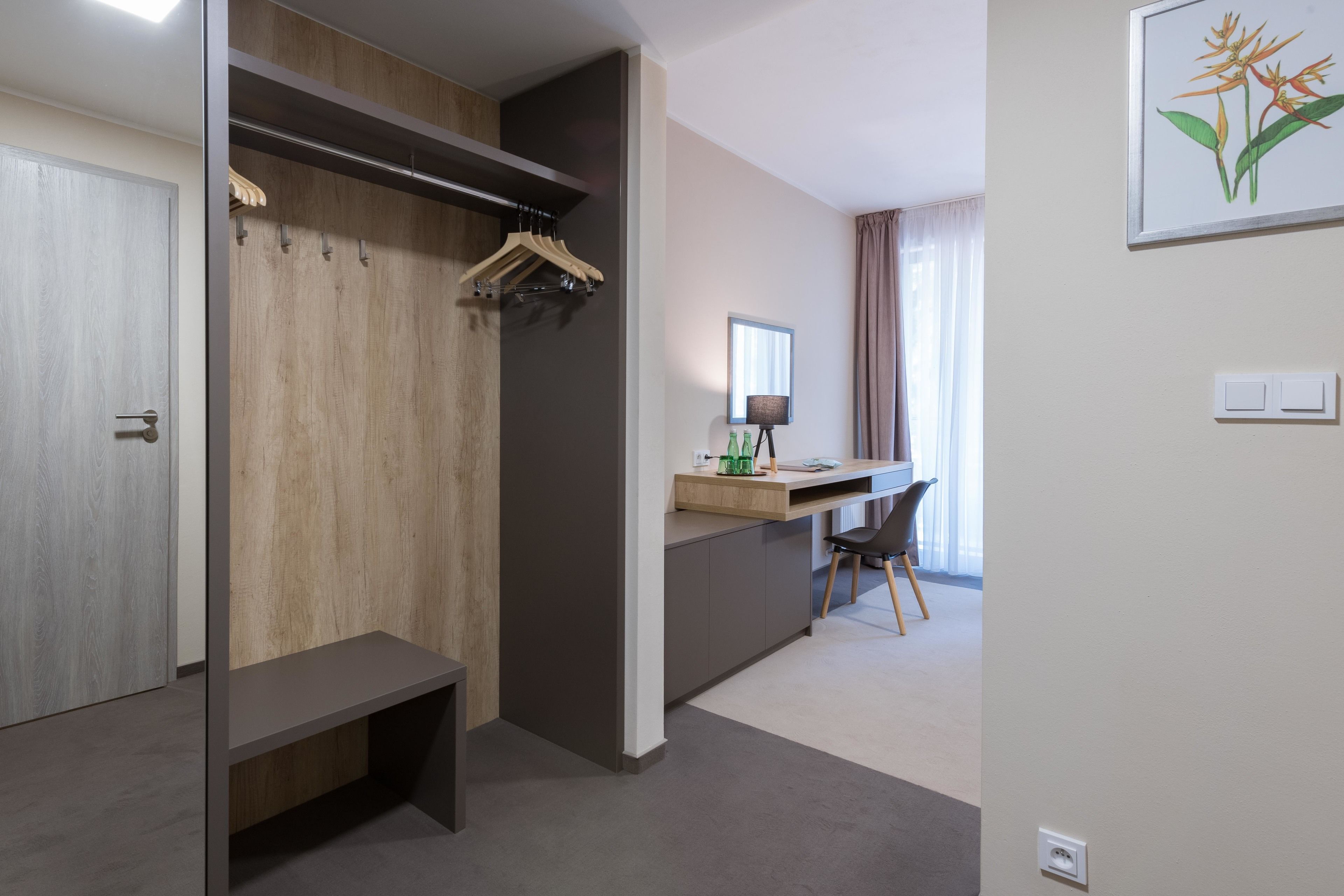 Een moderne hotelkamer met een houten garderobe, een bureau met stoel, en een klein bankje.
