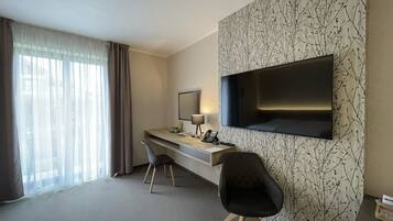 A hotel room with a desk, two chairs, a flat-screen TV mounted on the wall, and a large window with curtains.