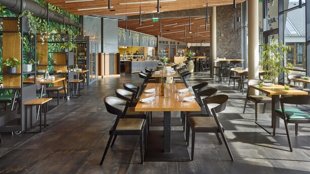 Een modern restaurant met een lange gedeelde tafel, losse zitplaatsen en een zichtbare keuken in de achtergrond.