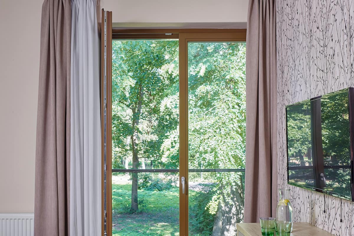 A bedroom with a bed, a desk with a chair, a window with curtains, and a view of greenery outside.