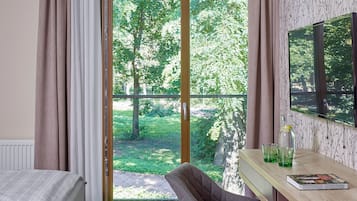 A bedroom with a bed, a desk with a chair, a window with curtains, and a view of greenery outside.
