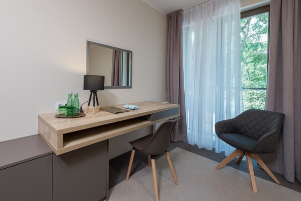 A modern room with a wooden desk, a black chair, a mirror, and a window with curtains. - Hotel Panská Zahrada (Dobrichovice)