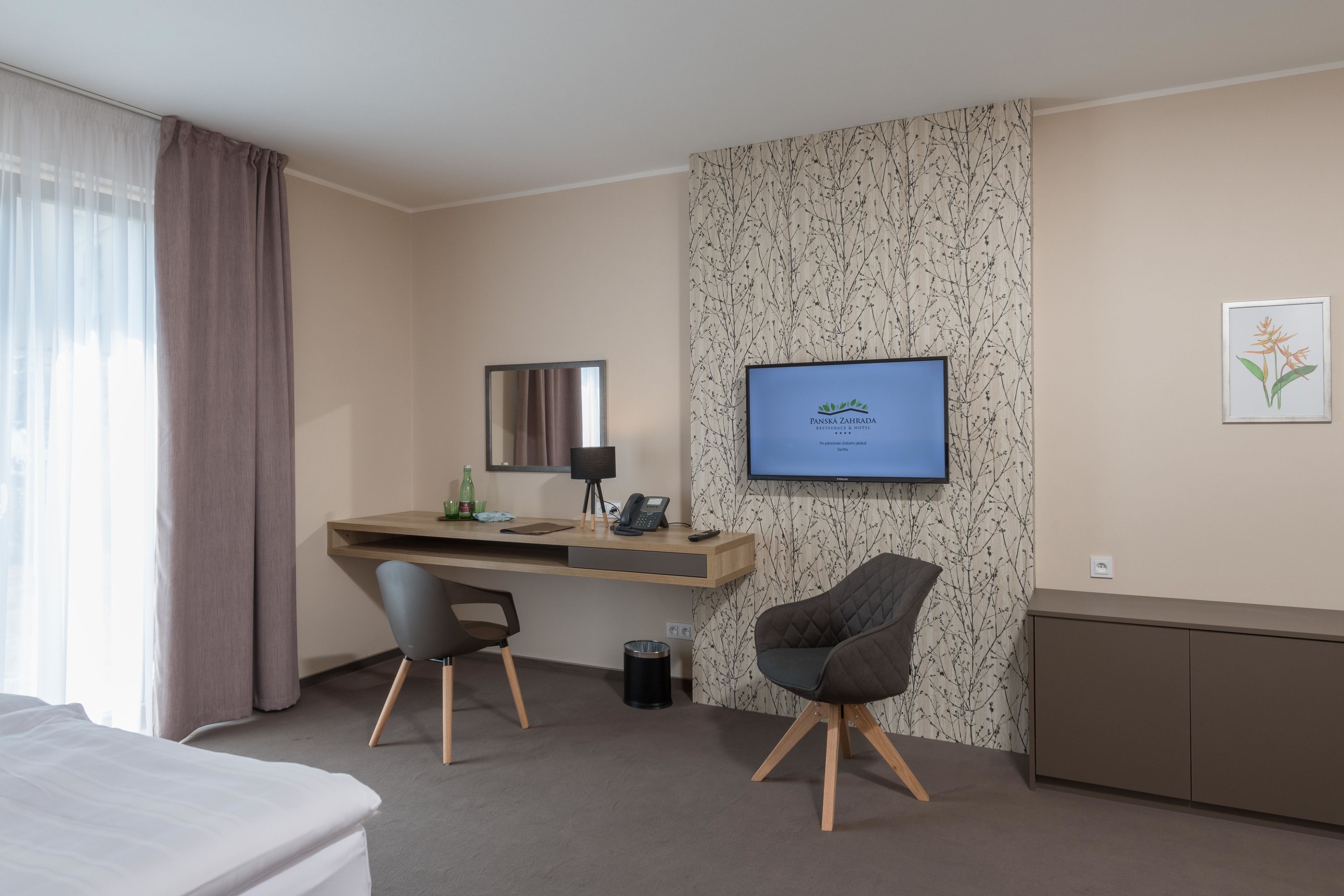 A hotel room with a bed, a desk with a computer, a chair, a TV, and a wall with a plant illustration.