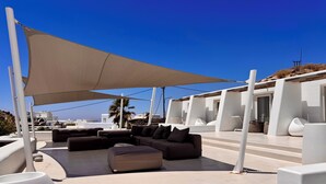 A modern outdoor lounge area with a large sectional sofa, a round table, and a canopy providing shade. - Amethyst Suite (Mykonos)