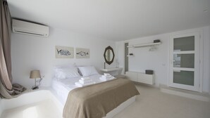 A bedroom with a bed, a bedside table, a lamp, a mirror, artwork, and a wardrobe. - Amethyst Suite (Mykonos)