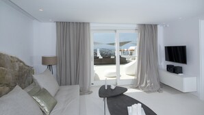 A modern living room with a sofa, a coffee table, and a TV. There is a balcony with a view of the city. - Amethyst Suite (Mykonos)