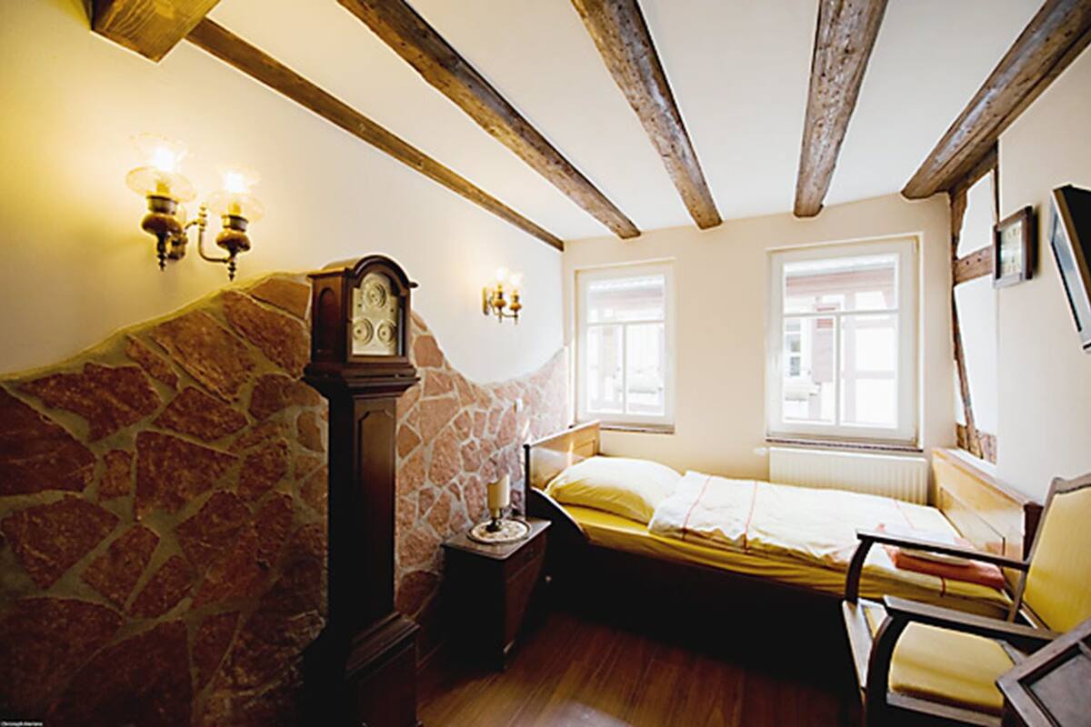 A bedroom with a stone wall, wooden beams, a bed, a nightstand, a clock, and two wall-mounted lamps.