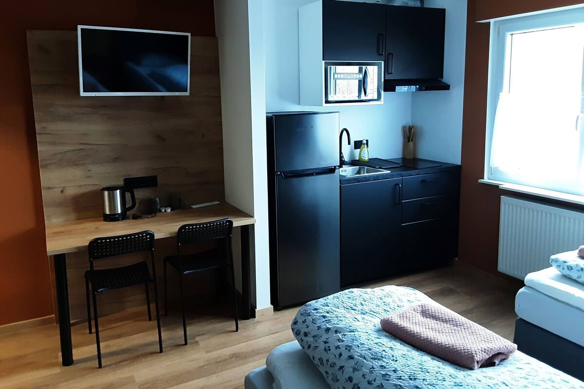 A compact room with a kitchenette, a bed with a patterned blanket, a TV mounted on the wall, and a window providing natural light.
