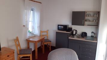 A compact kitchen with a microwave, kettle, and coffee maker on the counter, a small dining area with a table and chairs, and a cabinet with shelves above the sink.