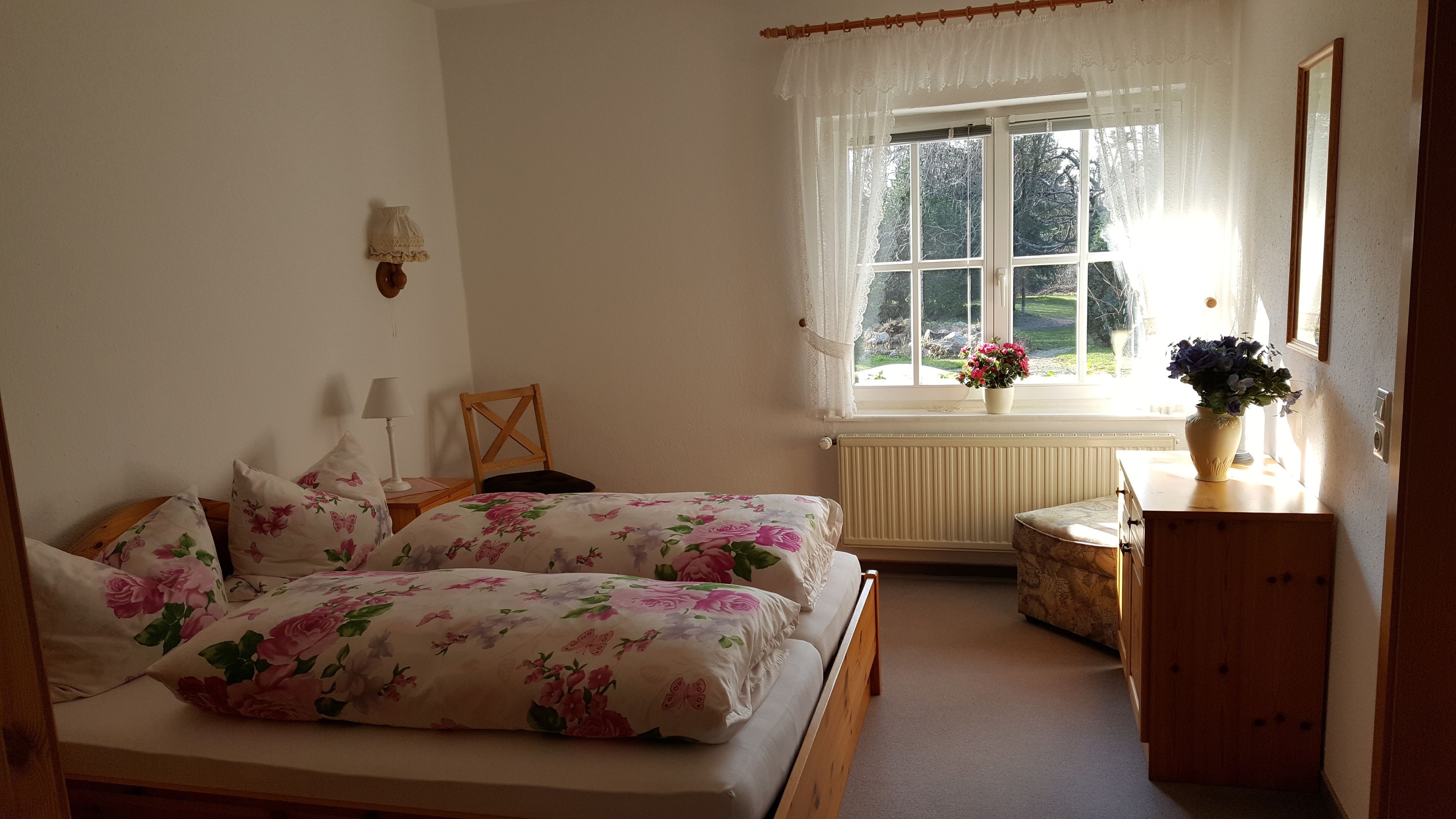 A bedroom with a double bed, a bedside table with a lamp, a chair, a dresser, a window with curtains, and a view of the outdoors.