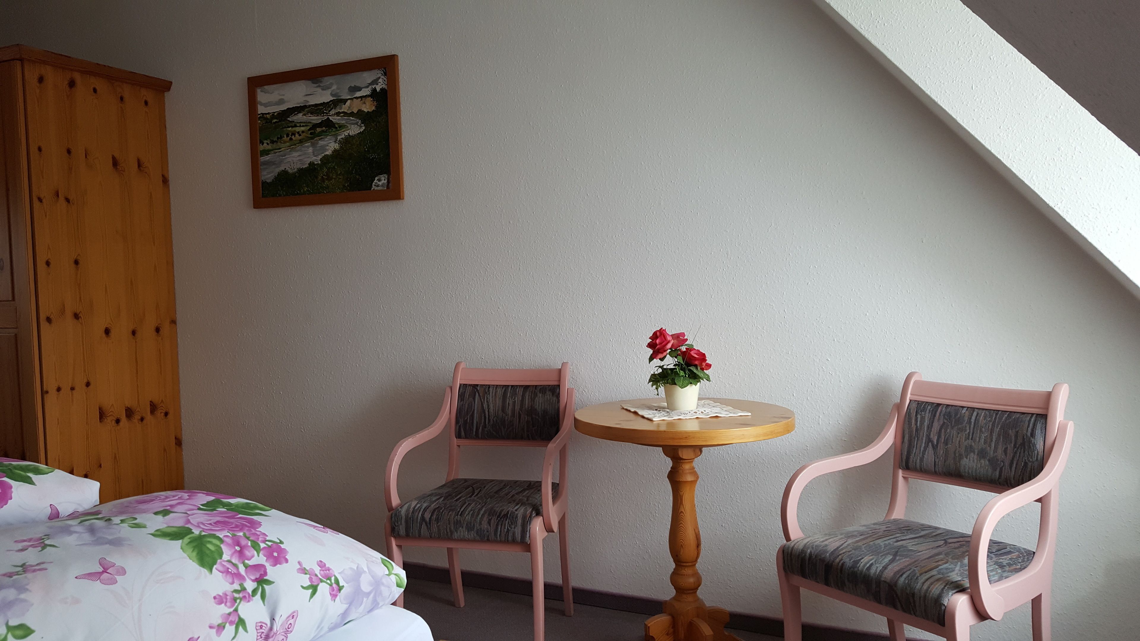 Una habitación con una cama, un armario de madera, un cuadro, dos sillas rosas, una pequeña mesa redonda con un jarrón de flores rojas y un techo inclinado.
