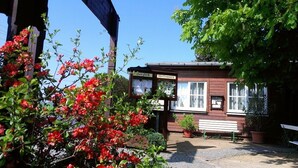 En liten, rustik byggnad med en skylt där det står ”Willkommen” (välkommen). Den har en träfasad, en bänk utanför och omges av frodig grönska och blommande växter.