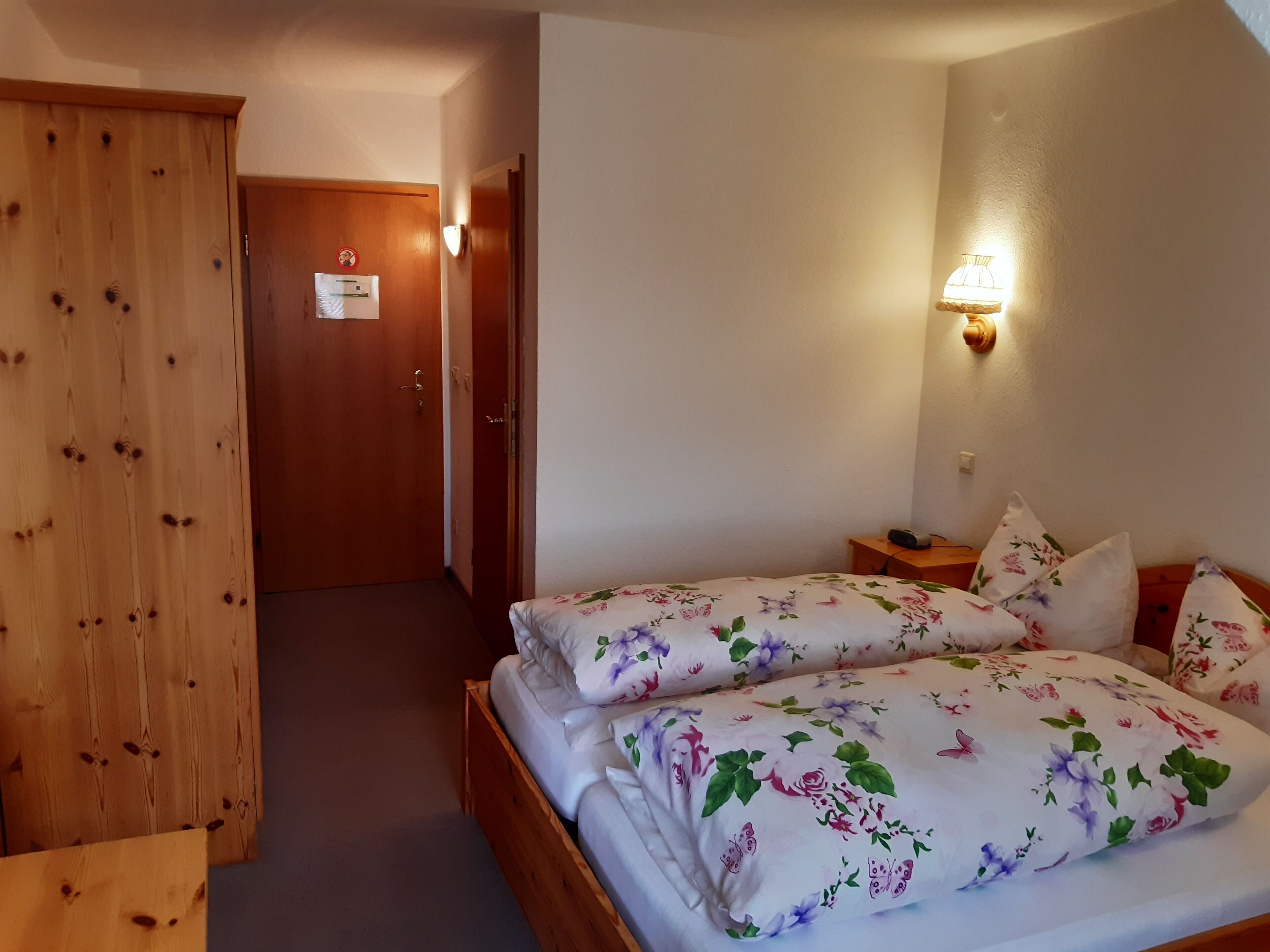 A hotel room with a wooden wardrobe, a bed with floral bedding, a bedside table with a lamp, and a door with a sign.