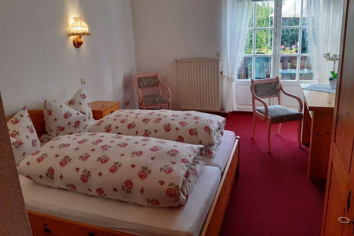 A bedroom with two beds, a small table, a chair, a window with curtains, and a radiator.