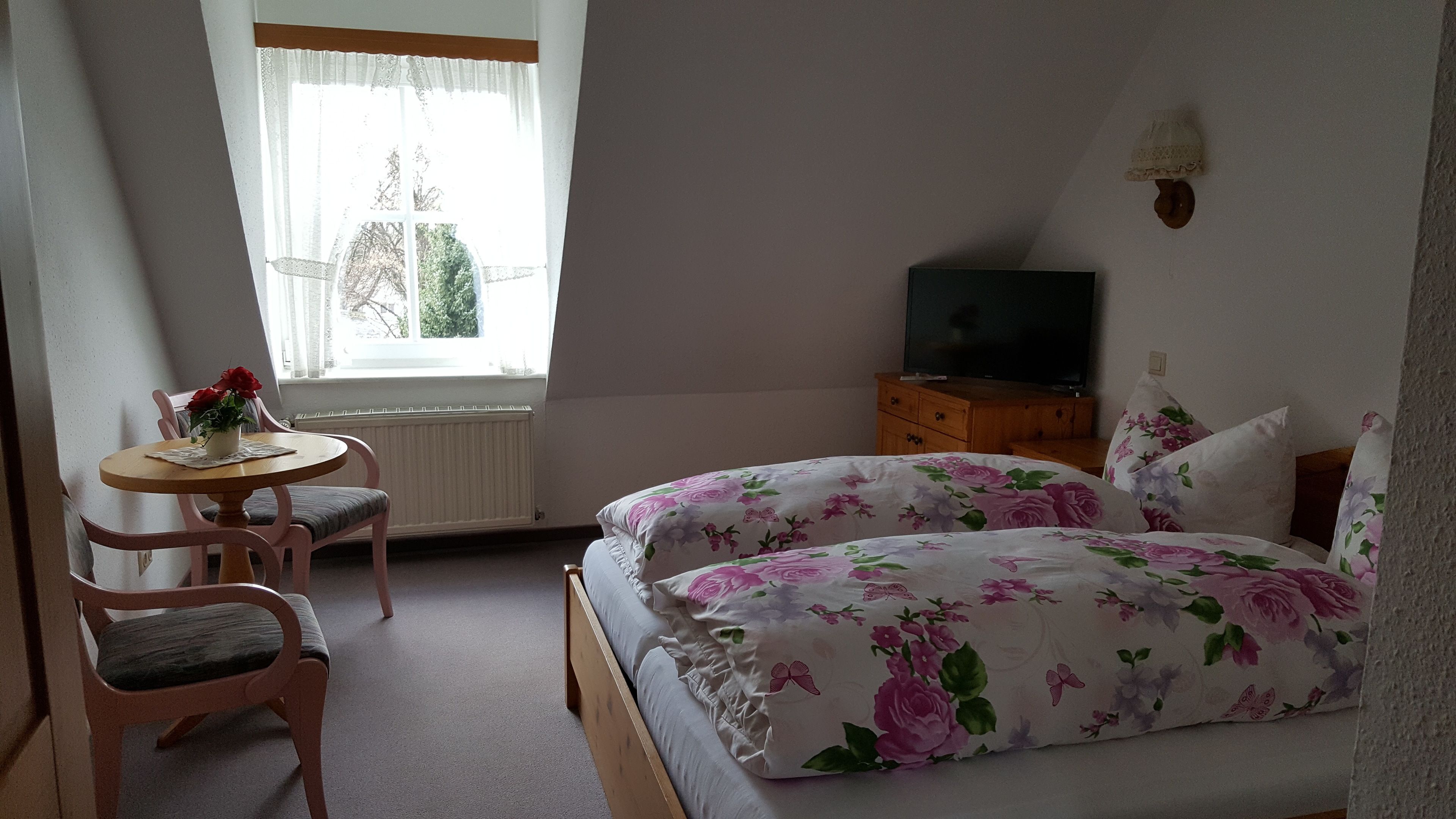 Ein Schlafzimmer mit einem Bett, einem kleinen Tisch mit Blumen in einer Vase, einem Sessel, einem Schminktisch und einem Fernseher.