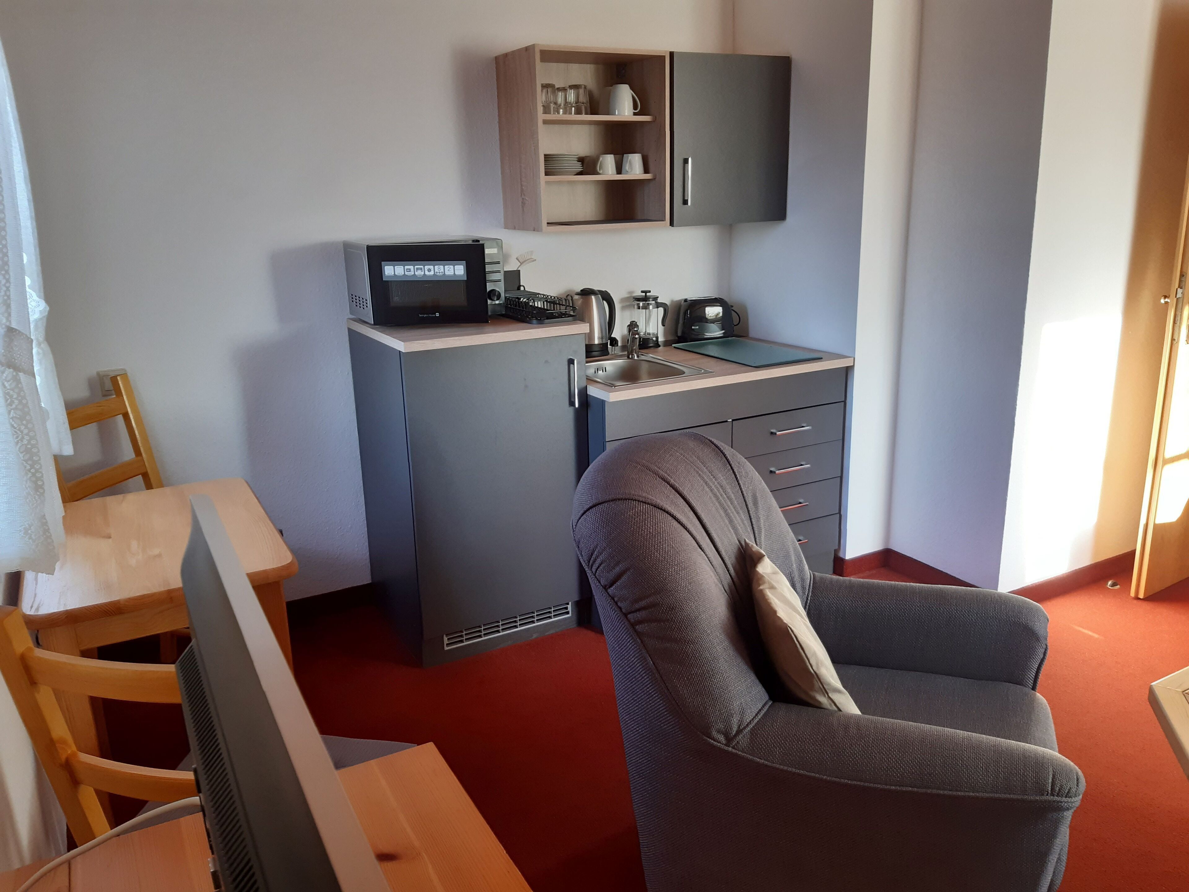 A compact kitchenette with a microwave, kettle, and sink. There is a chair and a small table in the room.