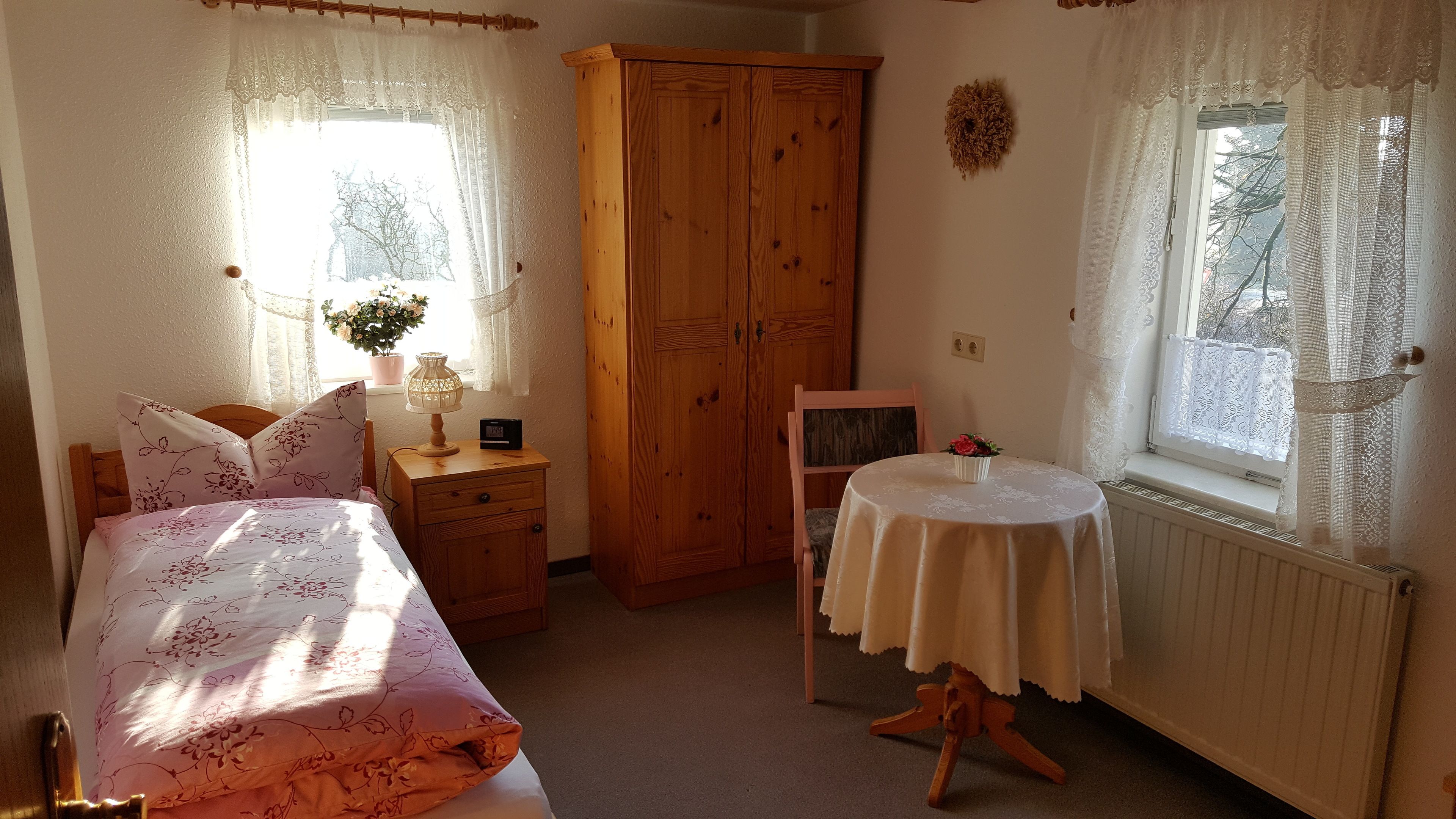A bedroom with a bed, a nightstand, a wooden wardrobe, a small table with a chair, and a window with curtains.