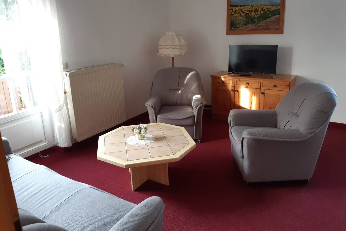 A living room with a sofa, two armchairs, a coffee table, a lamp, a TV on a wooden cabinet, and a painting on the wall.