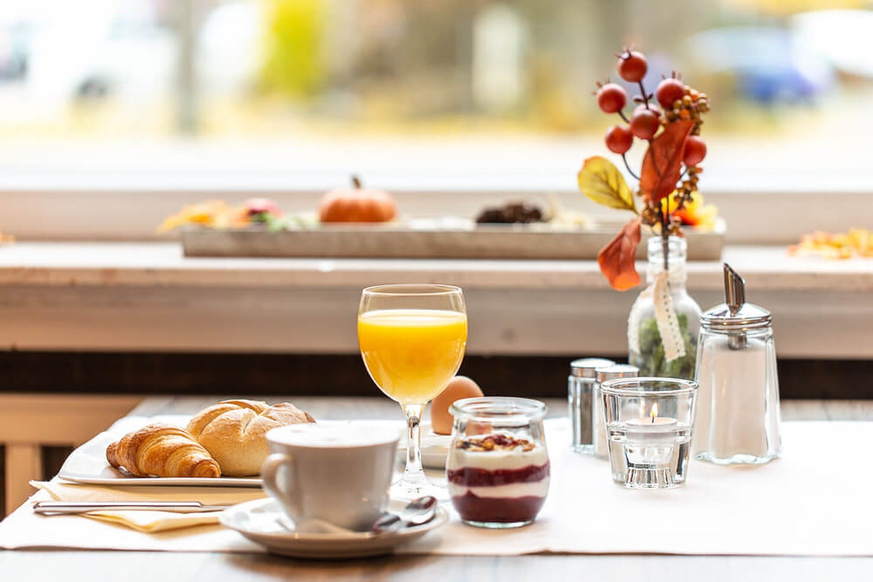 A breakfast setting with croissants, a glass of orange juice, a cup, a small jar of jam, and a vase with dried flowers.