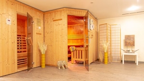 Un cuarto de sauna con paredes de madera y un banco. Hay una pequeña estatua en el suelo y una mesa con un florero de plantas secas.