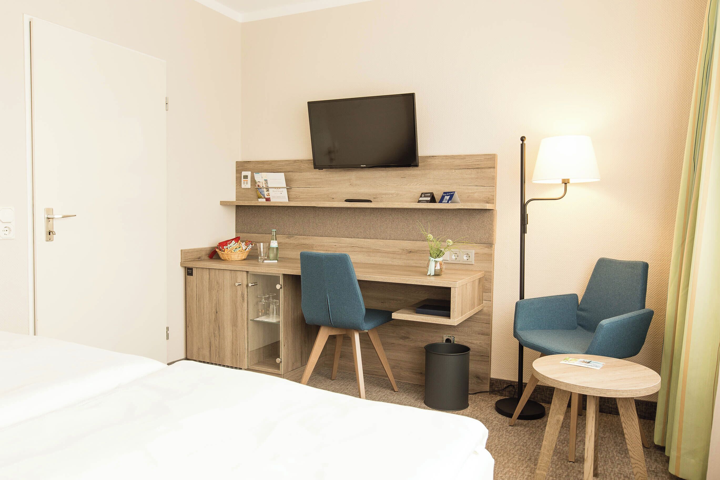 A hotel room with a bed, a desk with a chair, a television, a lamp, and a small table.