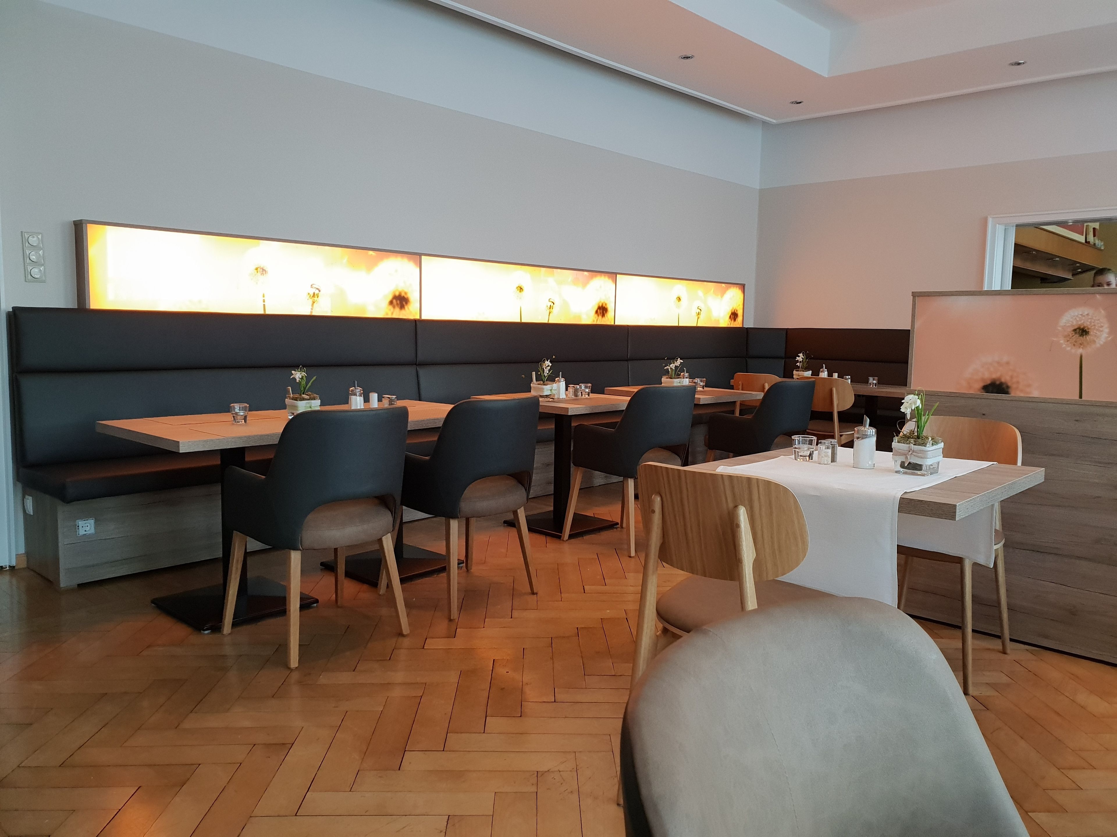 A modern restaurant with wooden tables and chairs, a black cushioned bench, and a wall-mounted light fixture.