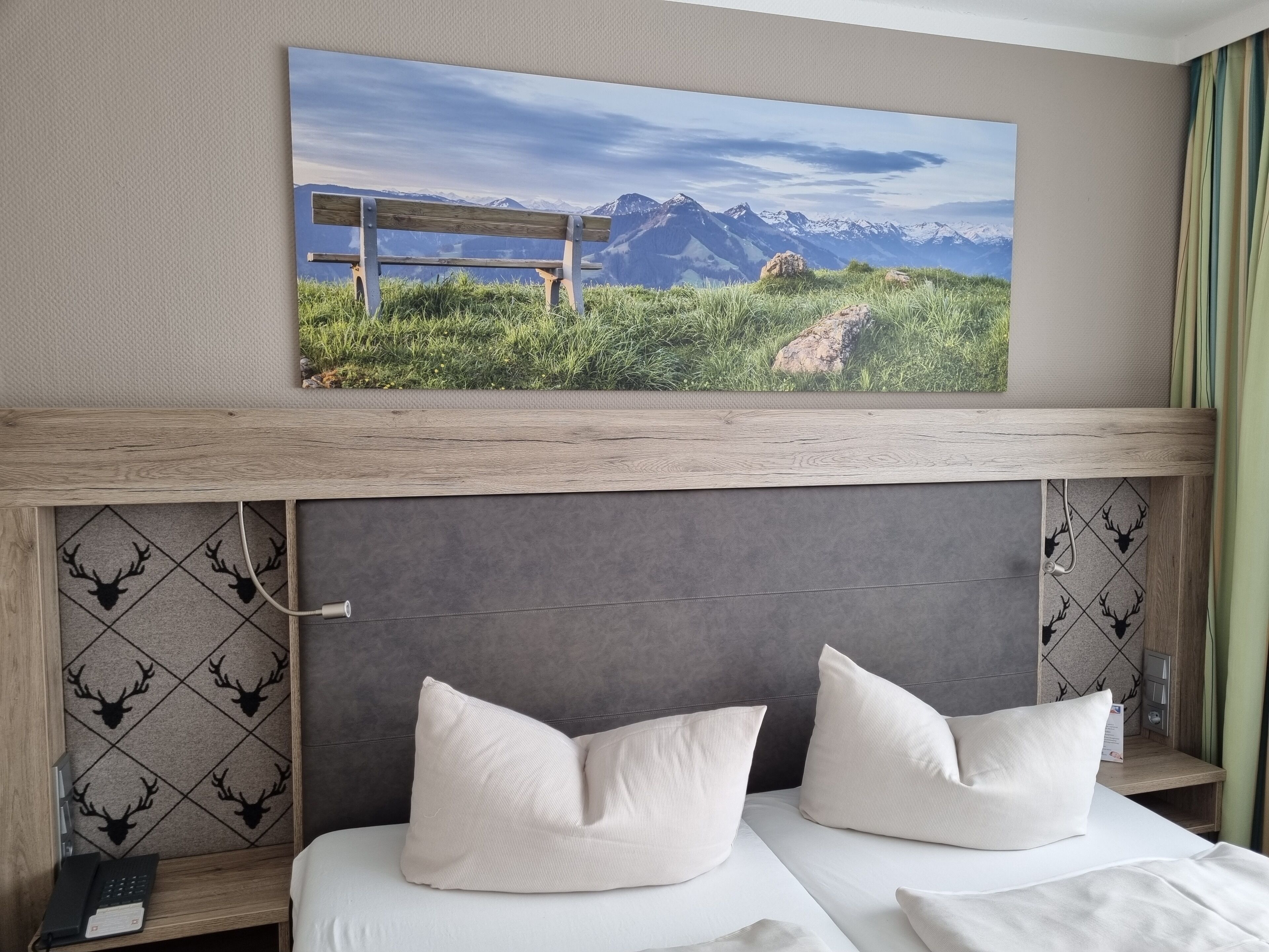 Habitación de hotel con cama, dos almohadas, un cuadro colgado de un paisaje montañoso y cabecera de madera.