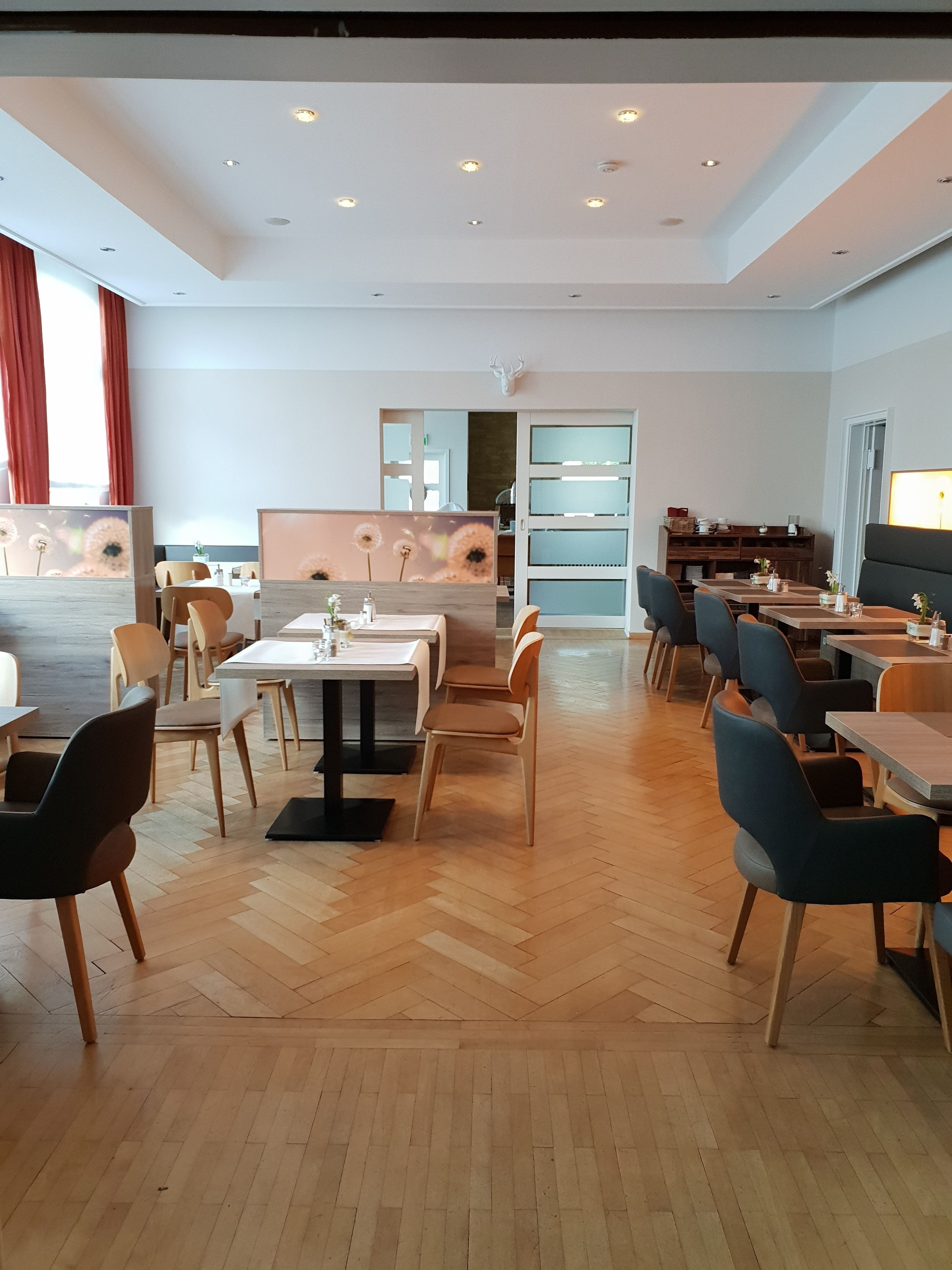 A modern dining area with wooden flooring, multiple tables and chairs, and a large window with red curtains.