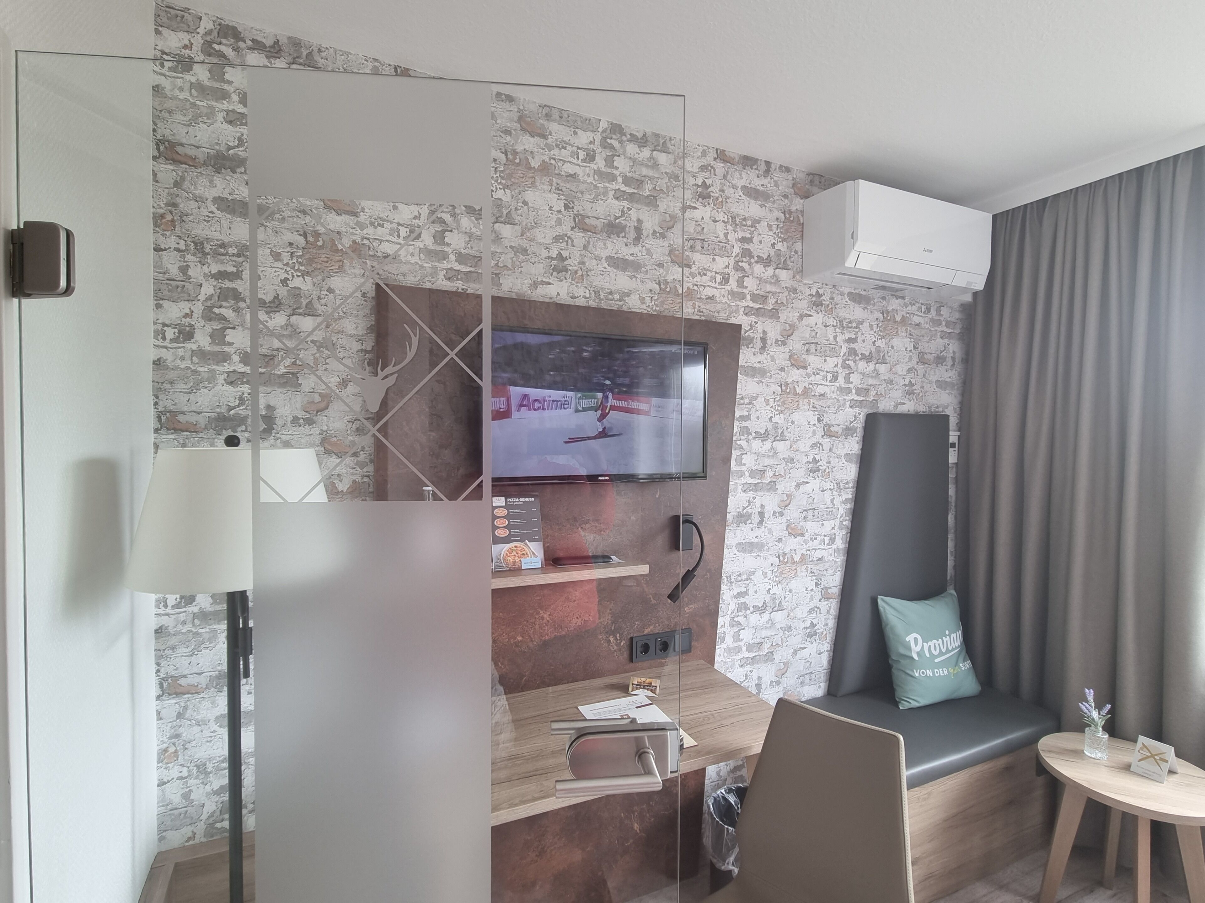 Une chambre d’hôtel moderne avec un mur au motif de briques, un téléviseur à écran plat fixé au-dessus d’un bureau en bois, un canapé gris avec un coussin et une petite table ronde sur laquelle est posé un vase.