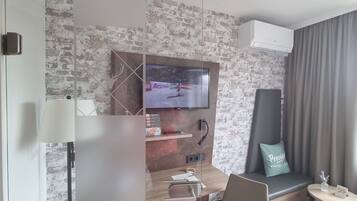 Une chambre d’hôtel moderne avec un mur au motif de briques, un téléviseur à écran plat fixé au-dessus d’un bureau en bois, un canapé gris avec un coussin et une petite table ronde sur laquelle est posé un vase.