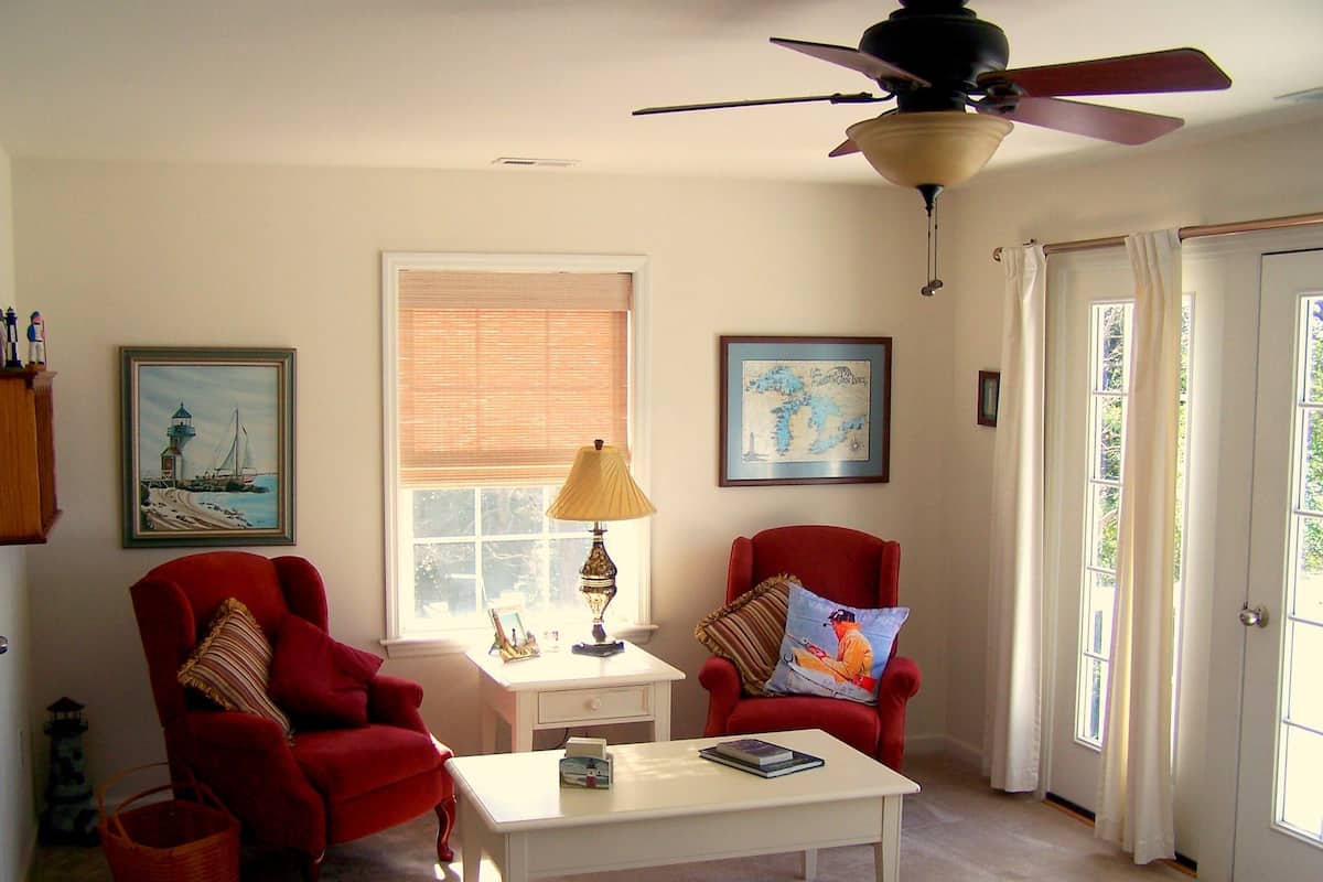 Un salon avec deux fauteuils rouges, une table basse blanche, une lampe, un ventilateur de plafond et une fenêtre avec des stores.