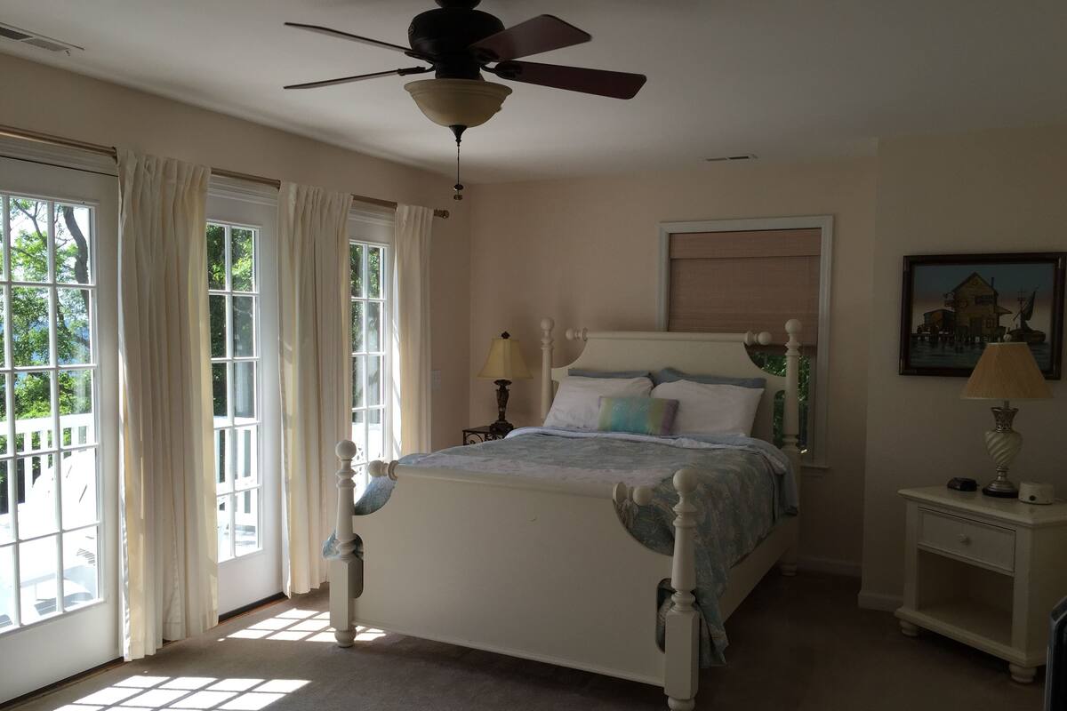 Une chambre à coucher avec un lit, un ventilateur de plafond, une table de chevet, une lampe et une fenêtre avec des rideaux.