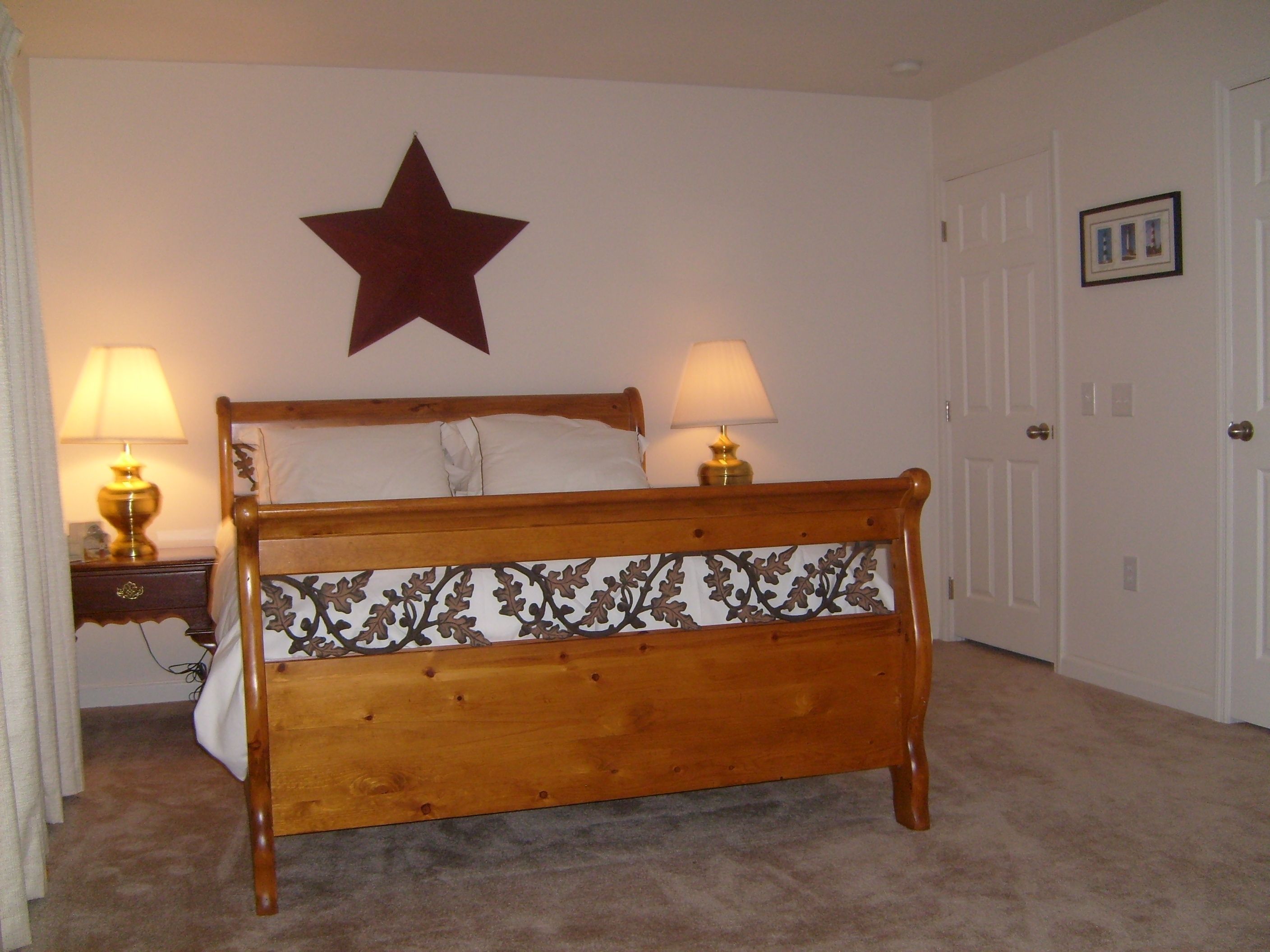 A wooden bed with a decorative headboard, two bedside lamps, a star decoration on the wall, and a door with a framed picture.