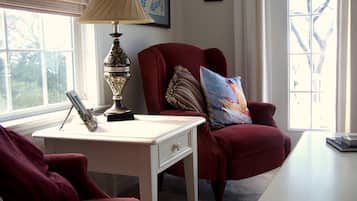 Una habitación con un sillón rojo, un escritorio blanco y una lámpara. Hay una ventana con persianas, un cuadro en la pared y una pequeña mesa con un libro y una lámpara.