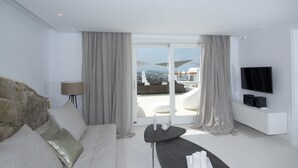 A modern hotel room with a stone accent wall, a sofa, a small table, a TV, and a balcony with a view. - Ruby Suite (Mykonos)