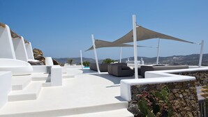 A sunlit outdoor seating area with white and gray lounge chairs, a stone wall, and a view of distant hills. - Ruby Suite (Mykonos)