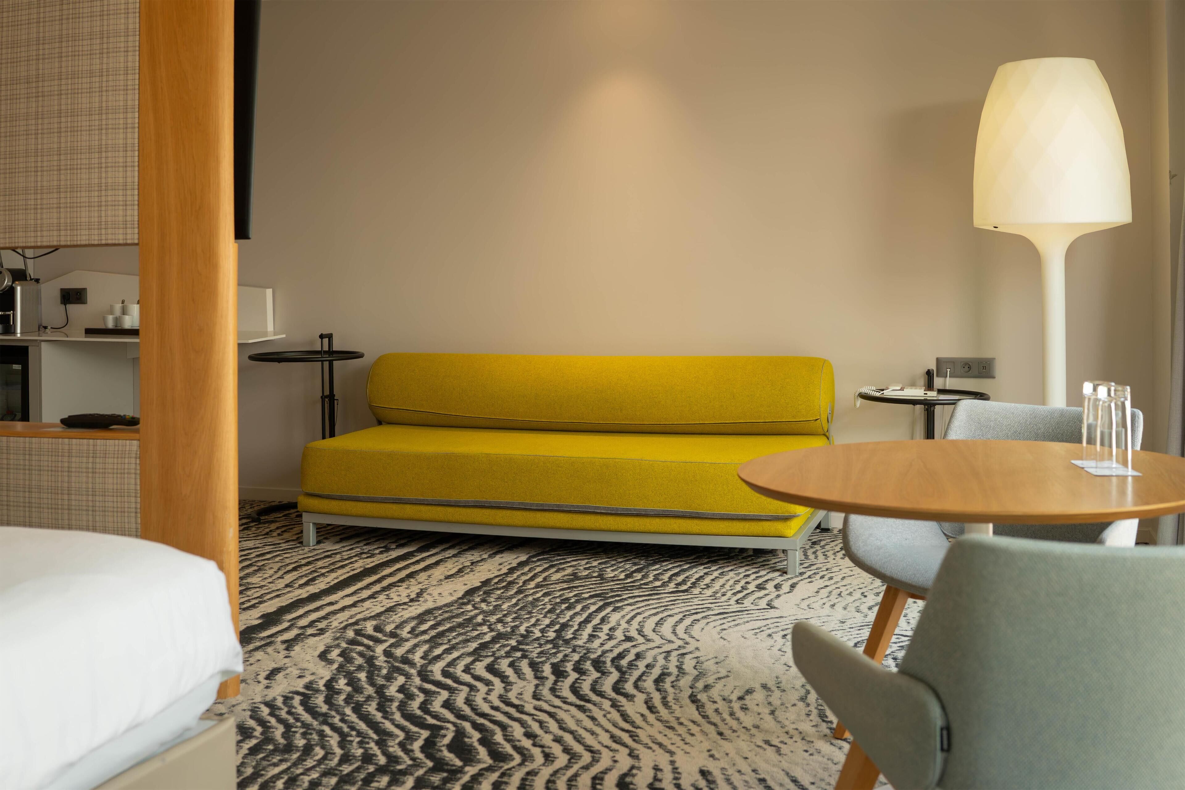 A hotel room with a bed, a yellow sofa, a round wooden table, and a lamp.