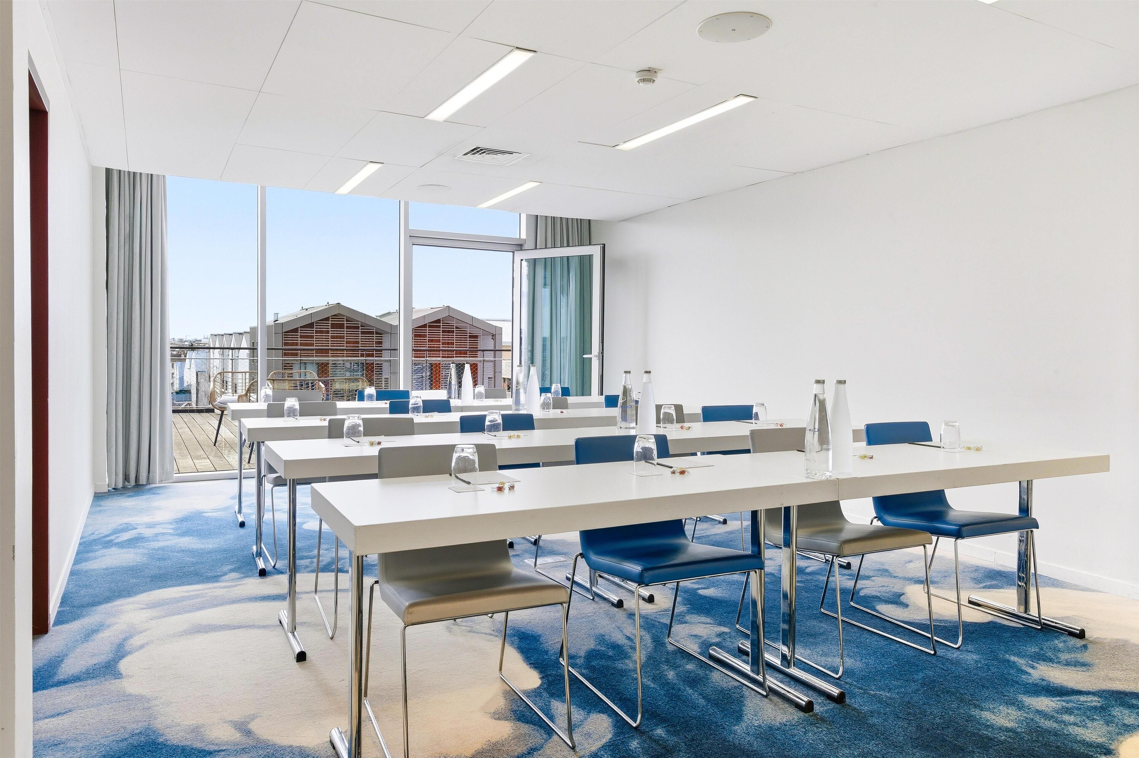 Un salón de conferencias con una larga mesa, sillas azules y vistas a edificios.