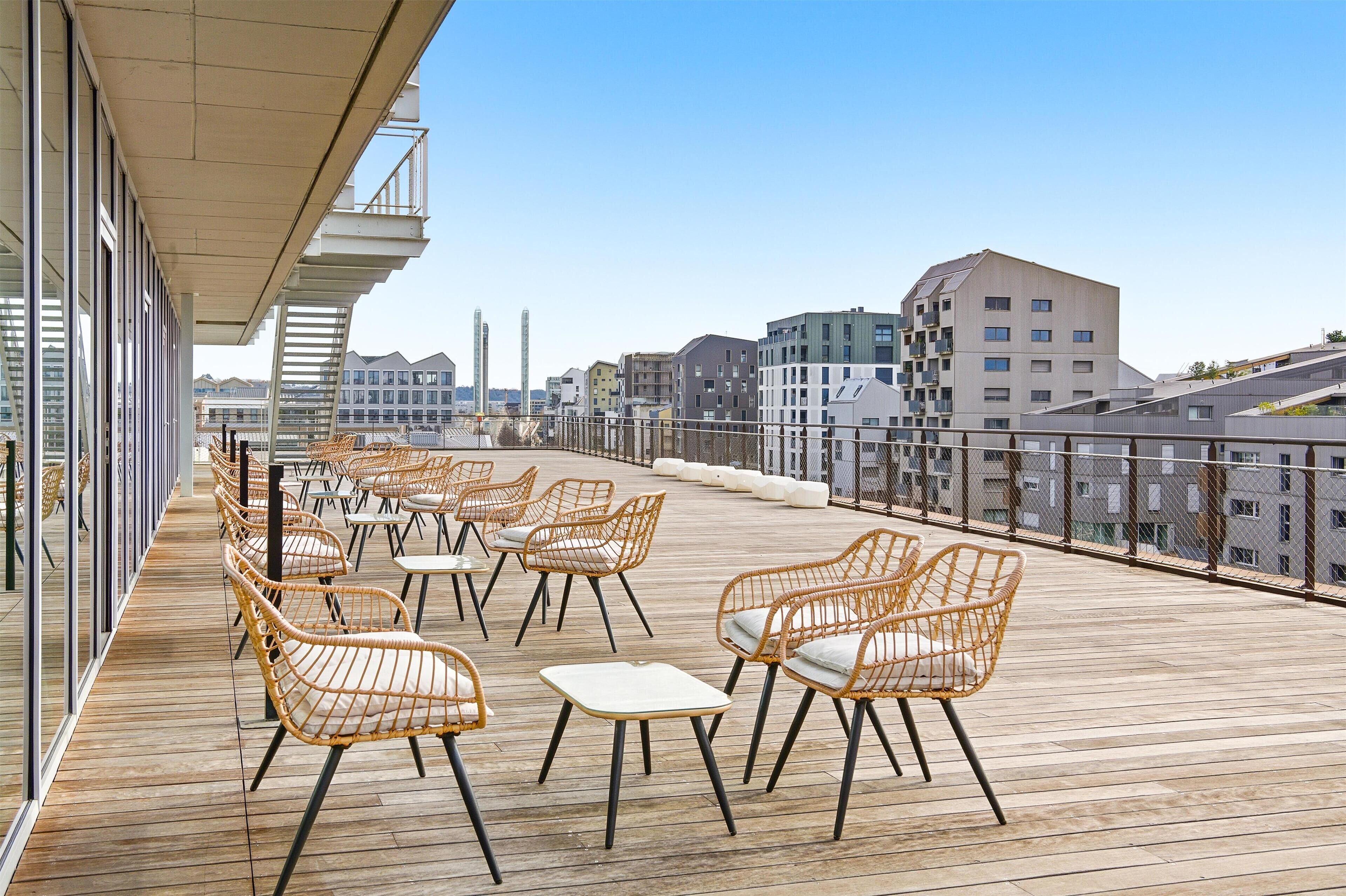 A rooftop terrace with wicker chairs and tables, overlooking a cityscape with modern buildings.