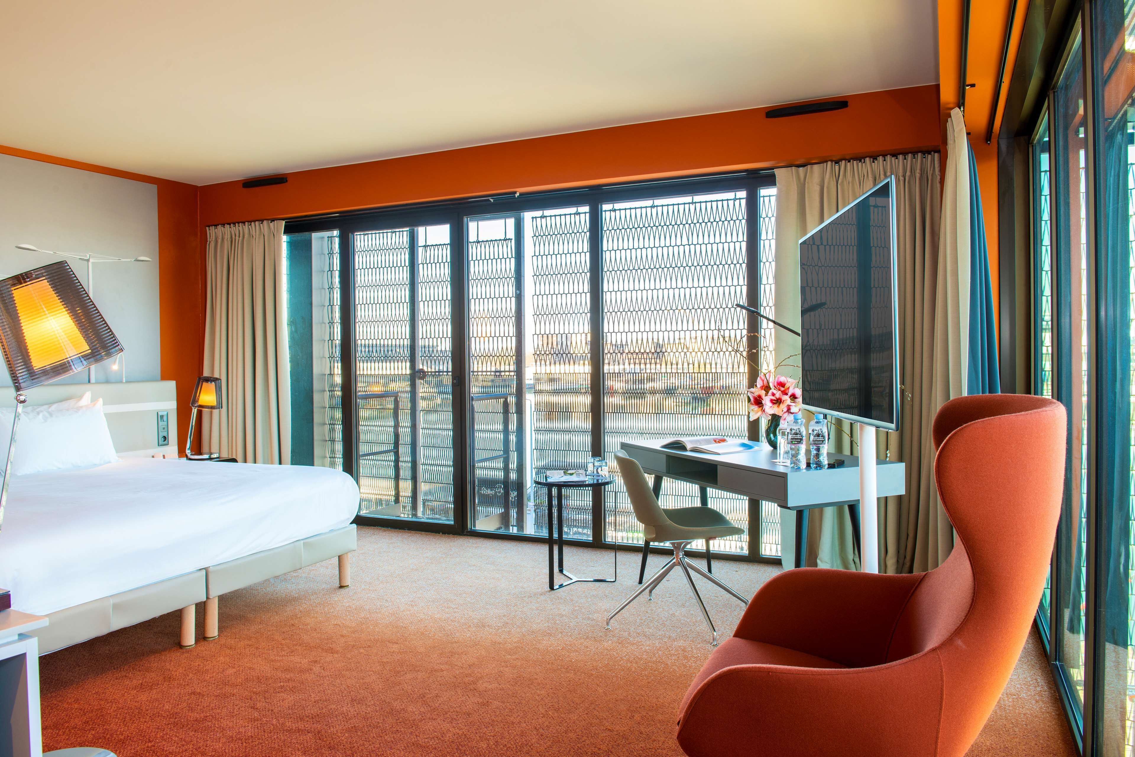 Habitación de hotel con cama, escritorio, silla, televisión y ventanas amplias.