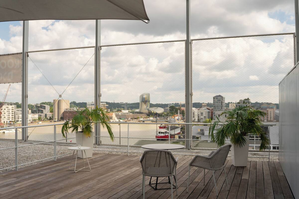 Un balcon avec deux chaises et deux plantes en pot, donnant sur un front de mer avec des bâtiments et un bateau rouge.