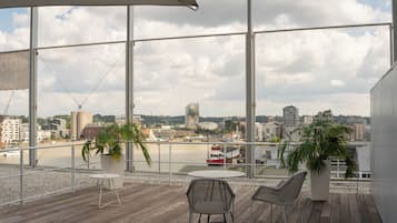 A balcony with two chairs and two potted plants, overlooking a waterfront with buildings and a red boat.