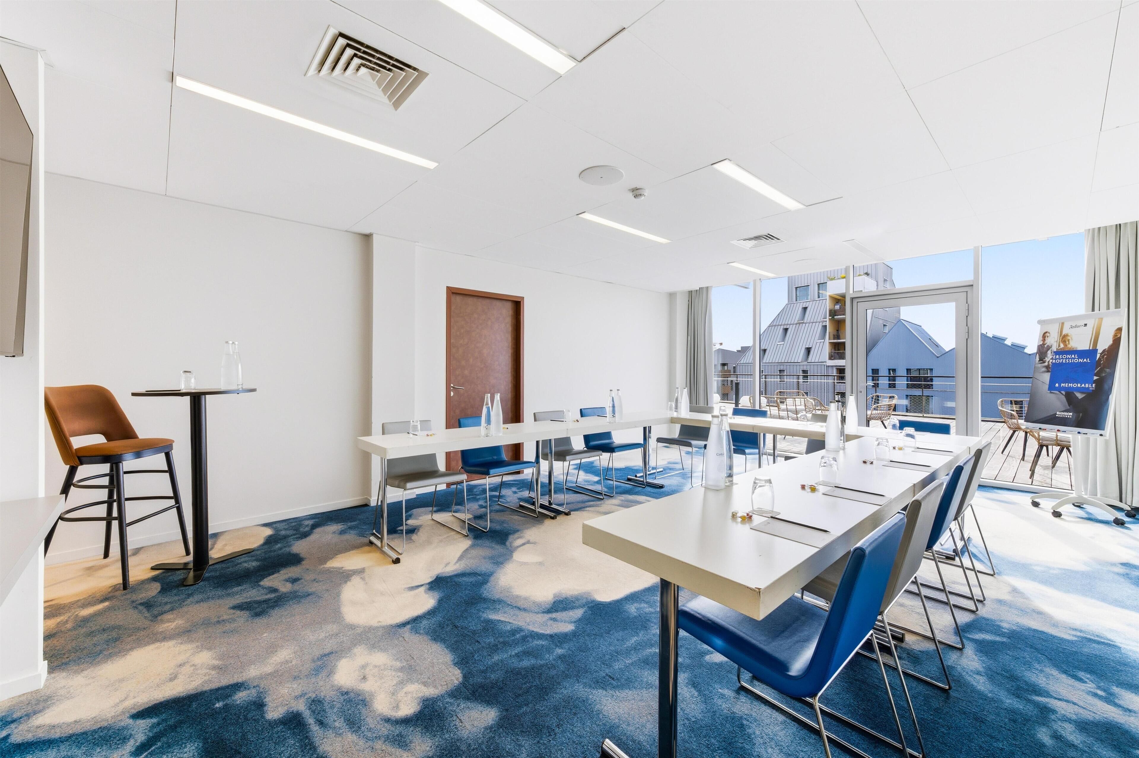 A modern conference room with a long table, chairs, and a view of buildings through large windows.