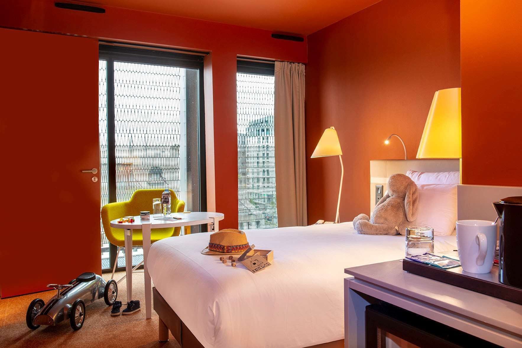 Ein Hotelzimmer mit Bett, Sessel, Tisch, Lampe, Teddybär und Spielzeugauto.