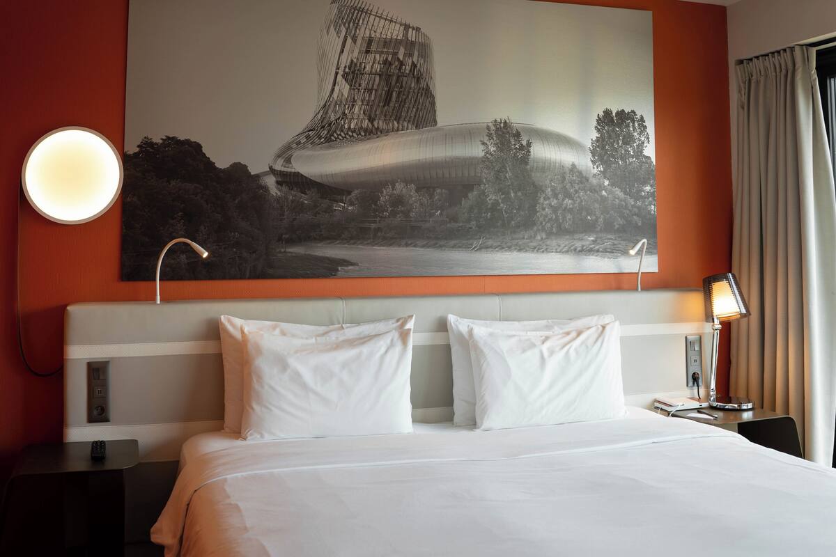 Une chambre d’hôtel avec un grand lit, deux tables de chevet, une petite table d’appoint, une lampe fixée au mur et un grand tableau encadré au-dessus du lit.